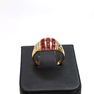 May include: A gold ring with a dome-shaped setting featuring a row of pink square-shaped stones surrounded by smaller clear stones.