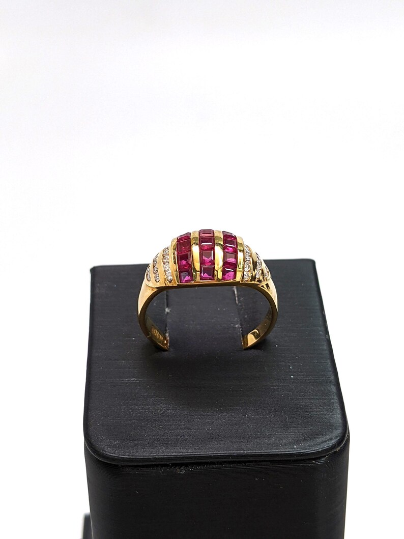 May include: A gold ring with a dome-shaped setting featuring a row of pink square-shaped stones surrounded by smaller clear stones.