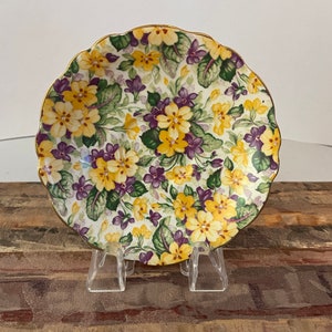 May include: A white saucer with a scalloped edge features a floral pattern of yellow and purple flowers on a green background. The saucer has a gold trim.