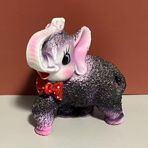 May include: A ceramic elephant figurine with a textured, speckled body in shades of purple and black. The elephant has pink ears, a white trunk, and a red bow tie with white polka dots. The figurine has a whimsical, vintage aesthetic.