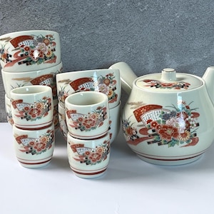 May include: A ceramic tea set featuring a teapot with a side handle and multiple small cups. Each piece is off-white with a decorative pattern of red, orange, and gold floral motifs, stylized fans, and thin red horizontal lines near the base.