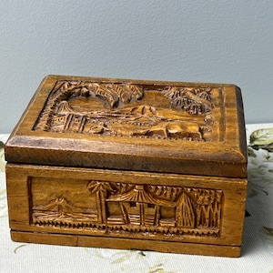 May include: A wooden box with intricate carvings of a tropical scene. The box is made of dark wood and has a hinged lid. The carvings depict palm trees, a hut, and a mountain.