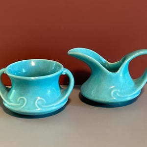 May include: A turquoise ceramic sugar bowl with two side handles and a matching turquoise ceramic creamer pitcher featuring a curved pouring spout and a single handle, both decorated with a raised swirl pattern at the base of the vessels.