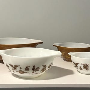 May include: Set of four vintage Pyrex mixing bowls. Two are white with brown decorative patterns, and two are brown. The bowls have a classic design with handles and are nested together.