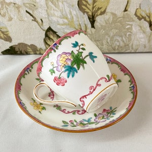 May include: A white teacup and saucer with a floral pattern in shades of pink, yellow, and green. The cup has a pink rim and a gold trim. The saucer has a pink rim and a gold trim.