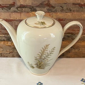 May include: White ceramic coffee pot with a gold trim and a floral design featuring brown ferns. The pot has a rounded body and a curved handle.