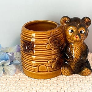 May include: A brown ceramic bear-shaped planter with a honey barrel design. The bear is sitting next to the barrel, which has a textured surface and floral accents. The planter is a decorative item.