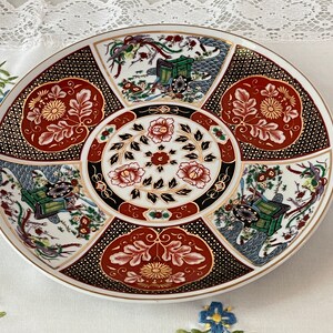 May include: A round, white ceramic plate with a gold rim. The plate is decorated with a colorful floral pattern and a central design of a cart pulled by a horse. The pattern includes red, gold, black, and blue colors.