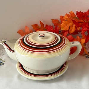 May include: A white ceramic teapot with a lid and a saucer. The teapot has a brown, orange, and white striped design. The saucer has a matching striped design.
