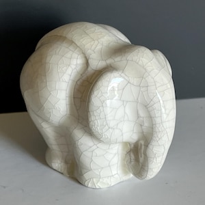May include: A ceramic elephant figurine featuring a glossy off-white glaze with a prominent dark crackle pattern across its entire surface. The sculpture depicts the animal in a standing position with its trunk curved downward toward the ground.