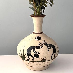 May include: A beige ceramic vase with a narrow neck and a rounded body. The vase features black Kokopelli figures, a crescent moon, and plant illustrations. The vase is filled with green and brown faux foliage.