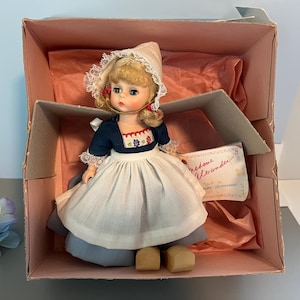 May include: A vintage doll in a pink box. The doll has blonde hair, blue eyes, and is wearing a blue and white dress with a white bonnet. The doll is in a box with tissue paper and a tag that reads "madame Alexander".