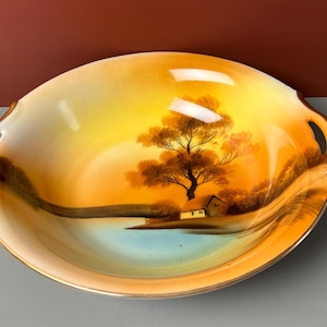 May include: Oval-shaped decorative serving dish with a landscape painting. The dish features a sunset scene with a tree, small house, and water. The rim and handles are gold-colored.