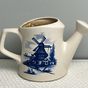 May include: White ceramic watering can with a blue and white windmill design. The windmill is a traditional Dutch design with a blue roof and white blades. The watering can has a spout and a handle.