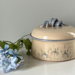 May include: A round, beige ceramic lidded container with a speckled finish, featuring a blue-tinted rim and a decorative, curled handle on the lid. The exterior is hand-painted with a pattern of blue and green iris flowers.