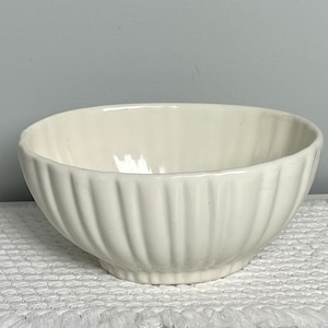 May include: Off-white ceramic bowl with vertical ribbed detailing. The bowl has a rounded shape and sits on a white surface. The bowl is empty and appears to be clean.