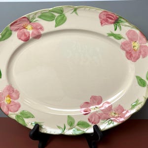 May include: Oval serving platter with a cream-colored base, adorned with pink floral and green leaf designs. The rim has a scalloped edge. The platter is supported by a black stand. A decorative piece for serving food.