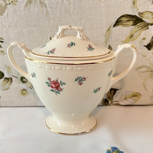 May include: White ceramic sugar bowl with a lid, featuring a floral pattern of pink roses and green leaves. The bowl has a gold trim around the top and base.
