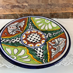May include: A large, round ceramic plate with a colorful, hand-painted design. The plate features a central circle with a black and white checkered pattern surrounded by green, yellow, white, and brown floral motifs.