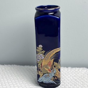 May include: A tall, rectangular, deep cobalt blue ceramic vase featuring a gold-rimmed opening. The lower section is decorated with a detailed illustration of a pheasant perched on a rock, surrounded by floral elements in gold, white, and green.
