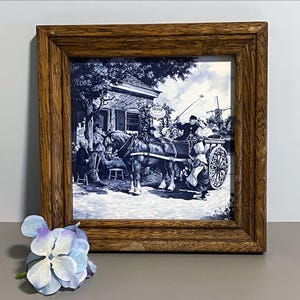 May include: A framed blue and white ceramic tile featuring a detailed scene of a horse-drawn carriage, people, and buildings. The tile is set in a brown wooden frame. A light blue flower is in the foreground.