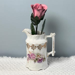 May include: A white porcelain pitcher with a floral design and gold accents, holding a pink metal rose with green leaves. The pitcher has a handle and a decorative spout, and is placed on a white, textured surface.