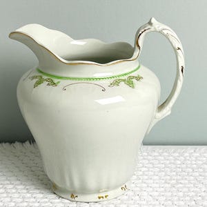 May include: A white ceramic pitcher featuring a scalloped rim with gold trim, a curved handle with gold accents, and a green decorative band around the upper body. The base of the pitcher is fluted and includes small gold painted details.