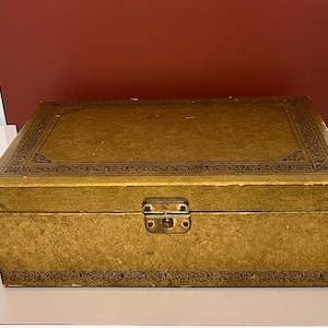May include: A rectangular gold-colored box with a hinged lid and a metal latch closure. The exterior features a textured finish and a dark brown decorative border pattern printed along the edges of the lid and the base of the box.