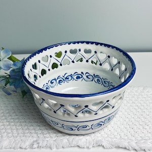 May include: White ceramic bowl with blue heart-shaped cutouts and decorative blue swirls. The rim and edges are painted blue. The bowl is on a white surface with blue flowers in the background. A decorative piece for the home.