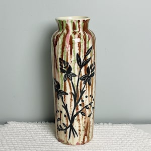 May include: A tall, cylindrical ceramic vase with a cream-colored base and vertical brown, green, and pink stripes. Black floral and leaf designs are painted on the front. The vase has a slightly flared top and is suitable for displaying flowers.