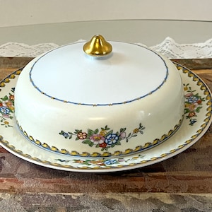 May include: A white porcelain butter dish with a floral design and a gold knob on the lid. The dish is on a wooden surface.