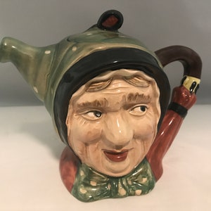 May include: A ceramic teapot shaped like a woman's head wearing a green and white polka dot hat. The teapot has a brown handle and a spout shaped like a woman's arm holding an umbrella.