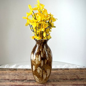 May include: A tall, decorative vase with a brown and white speckled pattern. The vase is filled with bright yellow flowers. The vase is on a wooden surface.