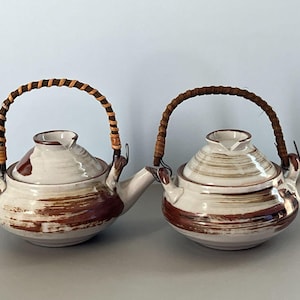 May include: Two ceramic teapots with lids and spouts. The teapots are off-white with brown brushstroke designs. Each teapot has a woven handle made of brown and tan material. The teapots are side by side.