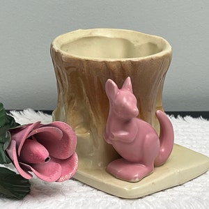 May include: A ceramic planter shaped like a tree stump with a pink kangaroo figurine attached to the side. The planter has a light brown glaze and the kangaroo has a light pink glaze.