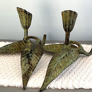 May include: Two bronze-colored candle holders. Each holder features a leaf-shaped base with a detailed vein pattern and a tulip-shaped cup for the candle. The holders have a weathered, antique appearance, adding a touch of vintage charm.