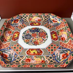May include: An octagonal decorative ceramic plate with a detailed, colorful design. The plate features a white center with a floral motif, surrounded by panels with birds, flowers, and geometric patterns in red, yellow, blue, and green.