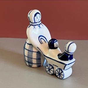 May include: A white ceramic figurine featuring a person in a bonnet pushing a small cart with two seated figures. The piece is decorated with hand-painted blue lines and a blue checkered pattern on the base, creating a simple, clean aesthetic.