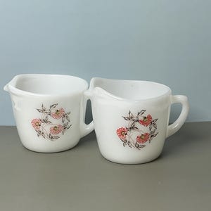 May include: Two white ceramic cream pitchers with a floral design. Each pitcher has a handle and a spout. The floral design features pink flowers and green leaves. The pitchers are on a gray surface against a blue background.