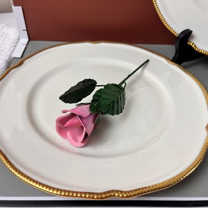 May include: A white ceramic plate with a gold beaded rim and scalloped edge. A pink metal rose with green leaves rests on the plate. The plate is displayed on a gray surface.