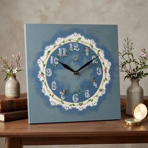 May include: Square wall clock with a blue background and a white, floral-decorated clock face. The clock hands are black, and the numbers are in a classic font. The clock is displayed on a wooden table with books and small vases.
