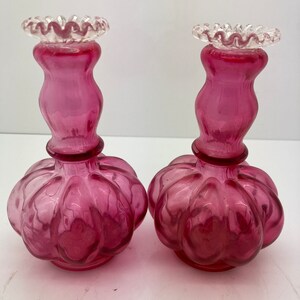 May include: A pair of pink glass candle holders with a ruffled top and a bulbous base.