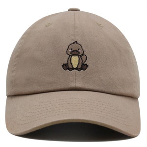 May include: A tan baseball cap with an embroidered cartoon platypus design. The platypus is brown with a yellow belly and has a pink nose and cheeks.