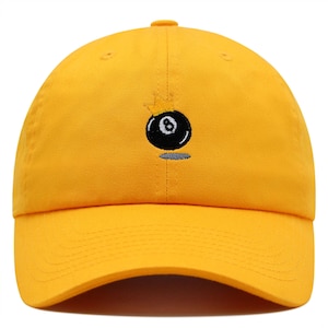 Könnte beinhalten: Eine gelbe Baseballkappe mit einem gestickten schwarzen 8-Ball mit einer goldenen Krone darauf.