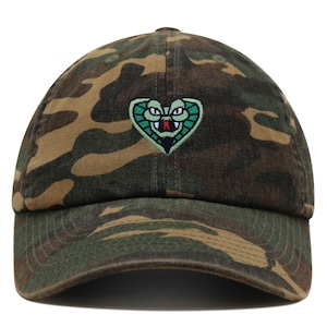 Peut inclure: Une casquette de baseball camouflage avec une tête de serpent verte brodée en forme de cœur. Le serpent a des yeux rouges et une bouche blanche.