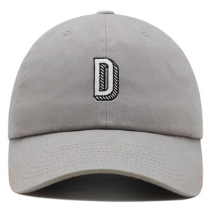 Puede incluir: Una gorra de béisbol gris claro con la letra D bordada en el panel frontal. La letra es blanca con un contorno negro grueso y un efecto tridimensional creado por líneas de sombreado diagonales negras en el perfil lateral de la letra.