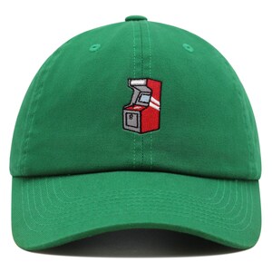 May include: A green baseball cap with an embroidered arcade game design. The arcade game is red, white, and gray.