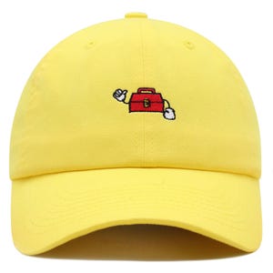 Puede incluir: Gorra de béisbol amarilla con visera curva. La gorra presenta una imagen bordada de dibujos animados de una caja de herramientas roja con guantes blancos y un gesto de pulgar hacia arriba. Hecha de tela suave y duradera.