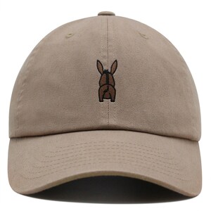 Puede incluir: Una gorra de béisbol de color beige con un burro marrón bordado en la parte posterior.