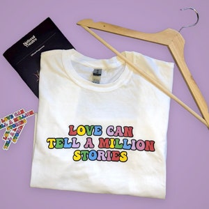 May include: White t-shirt with the text "LOVE CAN TELL A MILLION STORIES" in rainbow letters. The shirt is folded and on a purple surface with a wooden hanger and a National Theatre program.